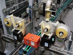 View of a system in which a carton is being labelled. A label dispenser is installed to the right and left of the conveyor belt.