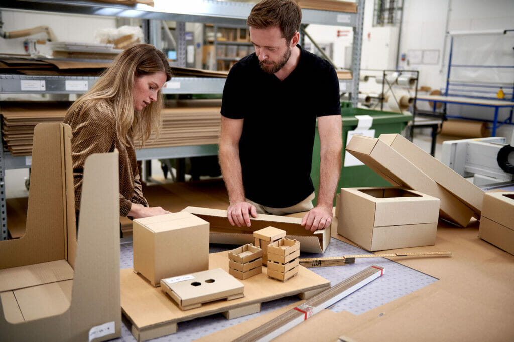 Ein Mann und eine Frau stehen an einem Tisch in einer Werkstatt, auf dem verschiedene Verpackungen und Verpackungselemente aus Pappe liegen.