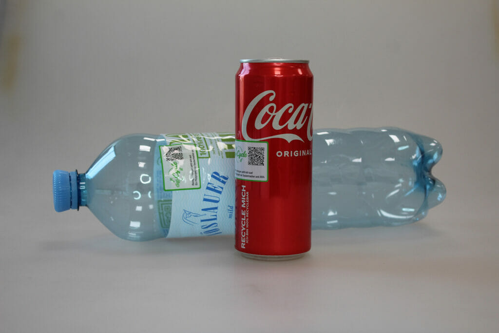 Hinter einer roten Cola-Dose liegt eine Plastikflasche. Beide Getränkeverpackungen sind mit einem Qr-Code beklebt.