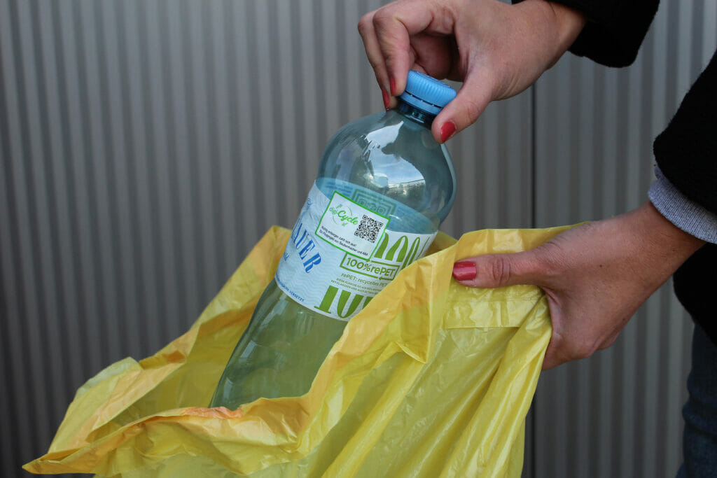 Nahaufnahme von zwei Händen. Die eine Hand hält einen Gelben Sack, in den die andere Hand eine Plastikflasche legt. Auf der Wasserflasche ist ein QR-Code vom Pilotprojekt "digi-Cycle" zu sehen.