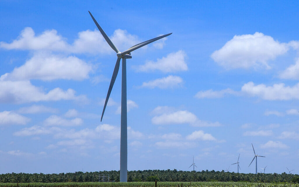 Vor blauem Himmel mit einzelnen Wolken steht ein Windkraftanlage auf einer Wiese und erzeugt erneuerbare Energie.