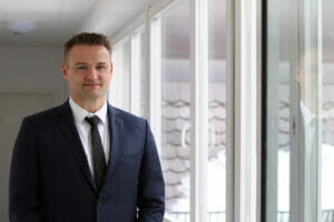 Foto von einem Mann im Anzug, der vor einer Fensterfront steht.