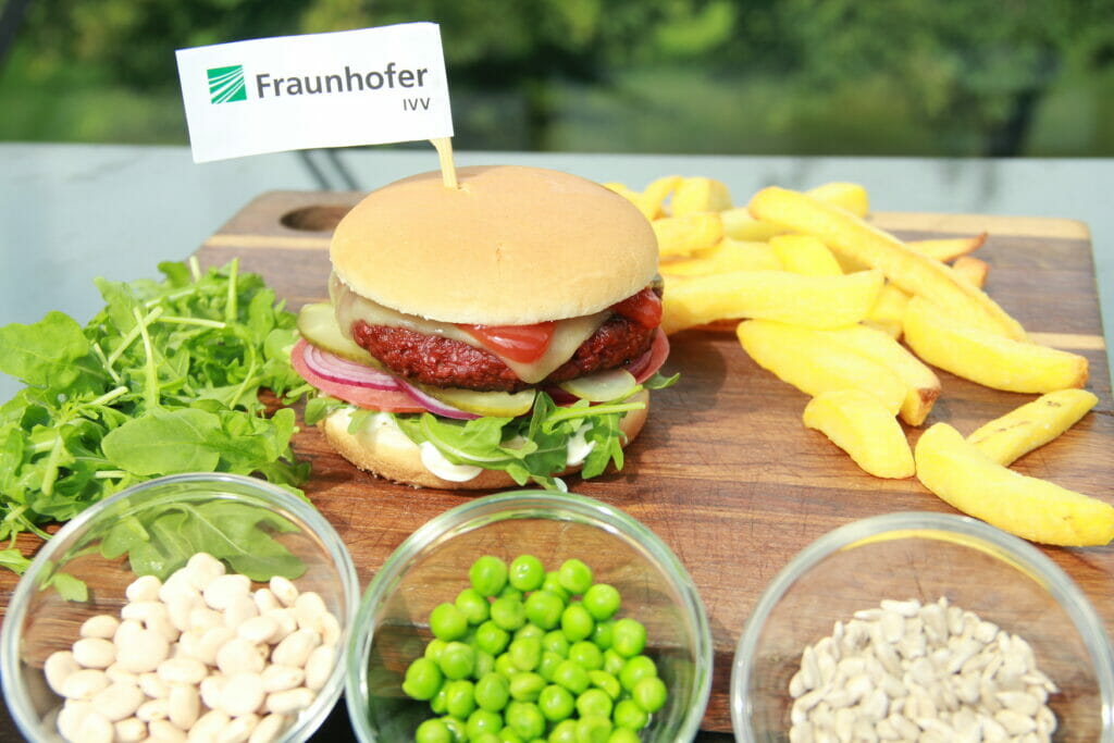 Auf einem Tisch liegen ein Burger, Pommes und Schalen mit Hülsenfrüchten.