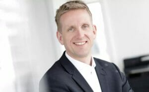Portraitfoto von lachendem Mann in schwarzem Anzug und weißem Hemd.
