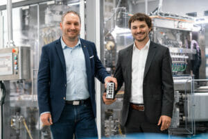 Zwei Männer in Anzügen stehen vor Maschinen.