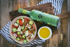 Auf einem Tisch stehen ein griechischer Salat, eine Schale mit Olivenöl und eine Flasche Olivenöl.