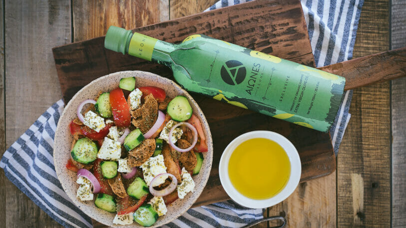 Auf einem Tisch stehen ein griechischer Salat, eine Schale mit Olivenöl und eine Flasche Olivenöl.