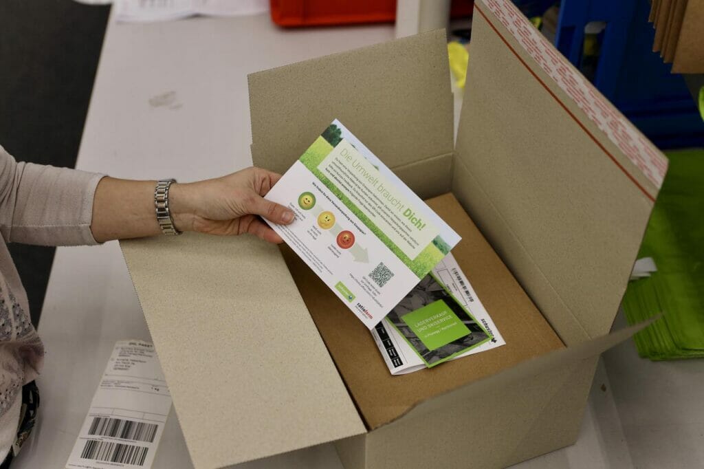 Bild von einem geöffneten Versandkarton. Eine Hand nimmt einen Flyer aus dem Karton.