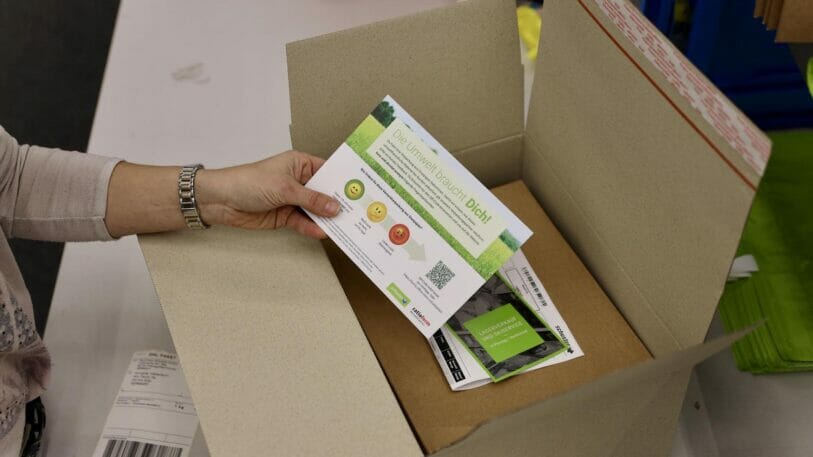 Bild von einem geöffneten Versandkarton. Eine Hand nimmt einen Flyer aus dem Karton.