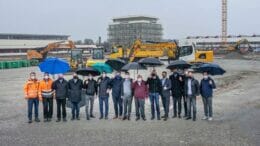Bild von einer Gruppe von Menschen mit Regenschirmen auf einer Baufläche, auf der ein Neubau entstehen soll.