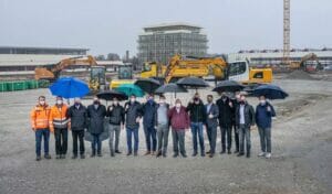 Bild von einer Gruppe von Menschen mit Regenschirmen auf einer Baufläche, auf der ein Neubau entstehen soll.