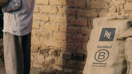 Bild von einer Ziegelwand von einem Haus, an der ein Sack mit dem Logo von Nespresso und der B-Corp-Zertifizierung lehnt. An der Wand geht eine Person vorbei.