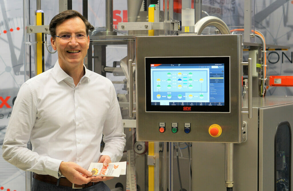 Picture of a smiling man holding packaged goods in his hands and standing in front of a packaging machine.
