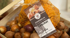 Bild von einem Sack mit Zwiebeln und einem Etikett, die auf losen Zwiebeln liegen.