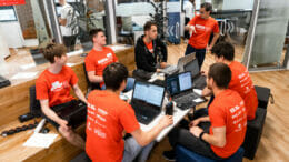 Bild von einer Gruppe von Menschen, die einer Person zuhören und an Laptops arbeiten