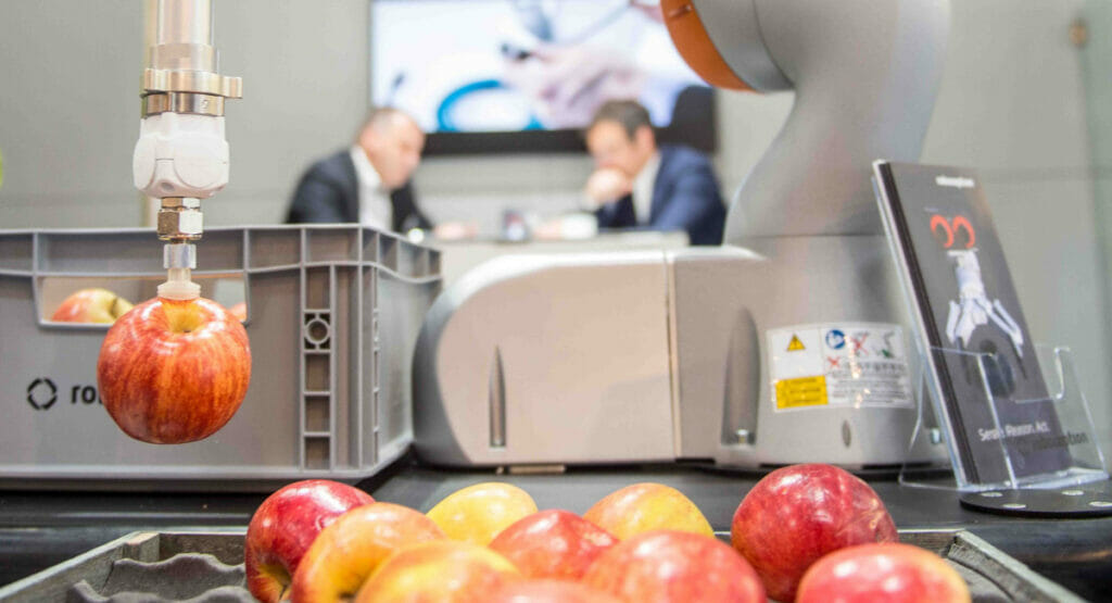 Bild von einem Roboter, der einen Apfel anhebt. Davor liegen weitere Äpfel und im Hintergrund sieht man zwei Männer, die sich unterhalten.