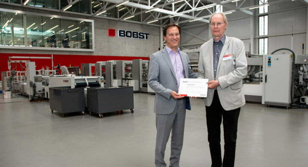 Bild von zwei Männern in einer Produktionshalle, die eine Urkunde in den Händen halten.