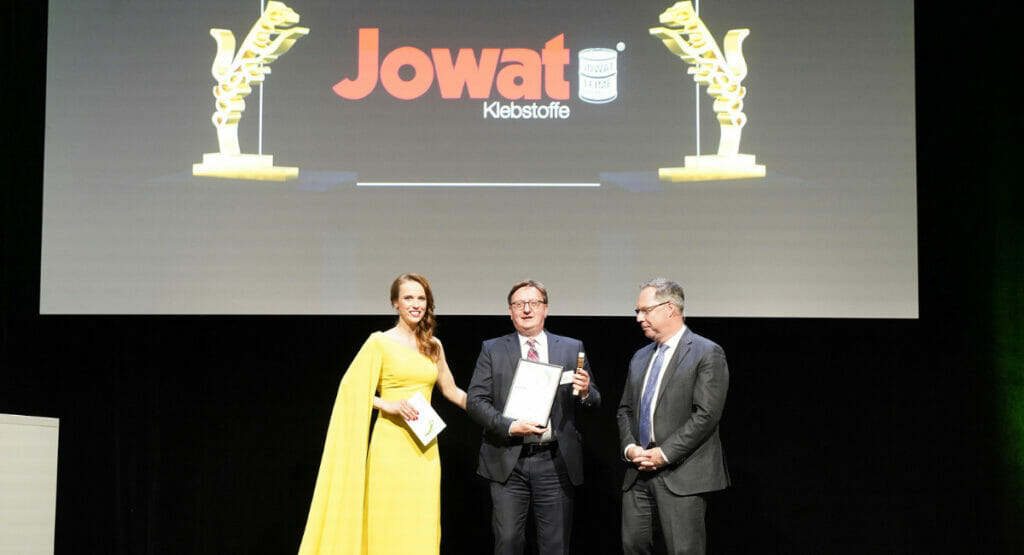 Bild von einer Bühne und einer Preisverleihung. Eine Frau mit zwei Männern.