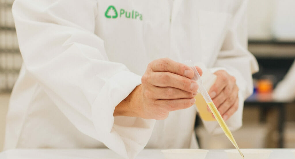 Bild von einer Person im Laborkittel, die eine Pipette mit Flüssigkeit in der Hand hält.