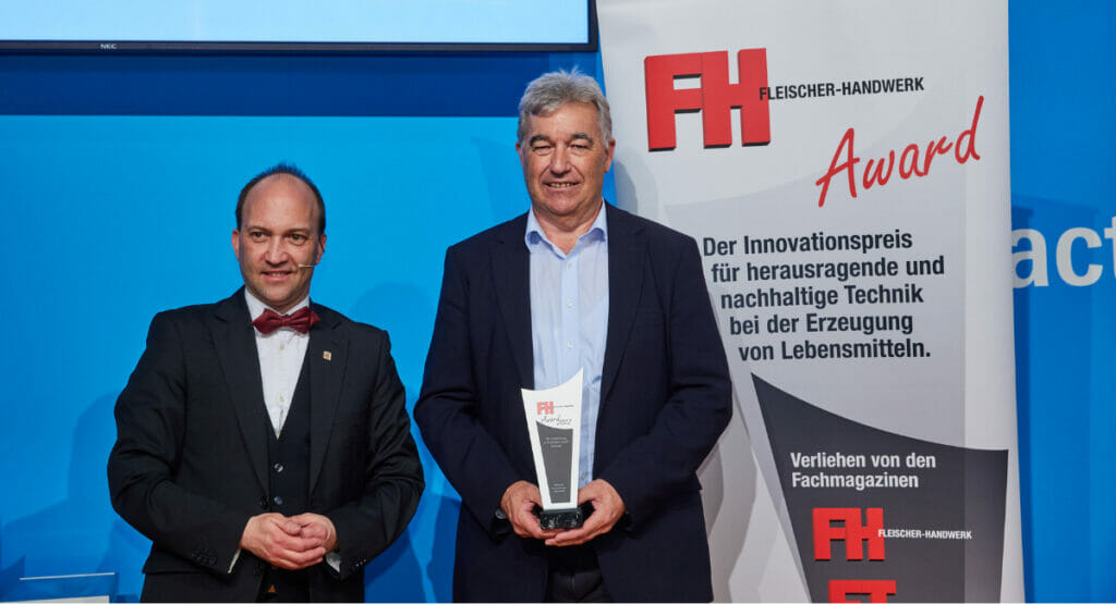 Bild von zwei Männern bei einer Preisverleihung. Der rechte Mann hält einen Preis in der Hand.