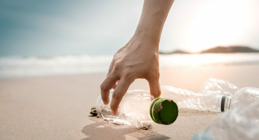 Bild von einer Hand, die an einem Strand eine Kunststoffflasche aufhebt.