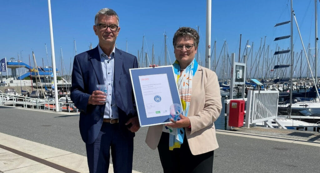 Bild von einem Mann und einer Frau, die an einem Hafen stehen und eine Urkunde und Becher halten.