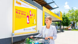 Bild von einer Frau mit Einkaufswagen, die an einem Aufklärungsplakat von Netto und der Initiative "Mülltrennung wirkt" zur richtigen Mülltrennung vorbeigeht.