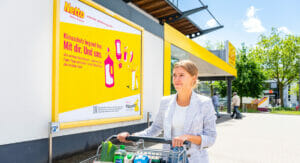 Bild von einer Frau mit Einkaufswagen, die an einem Aufklärungsplakat von Netto und der Initiative "Mülltrennung wirkt" zur richtigen Mülltrennung vorbeigeht.