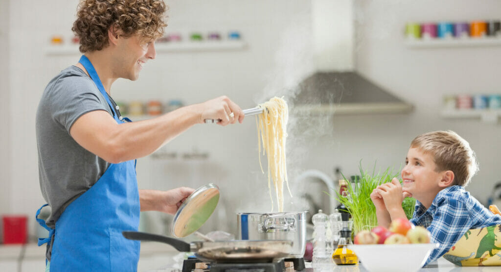 Bild von einem Mann, der in einer Küche steht und Pasta kocht und einem kleinen Jungen, der ihm dabei zusieht.