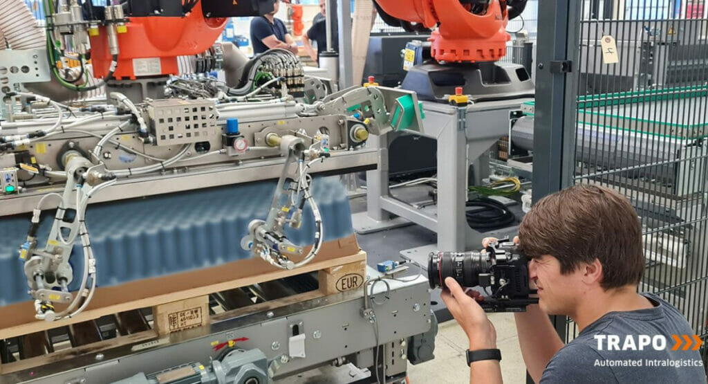 Bild von einem Mann, der vor einer Palettierungsanlage kniet und ein Bild mit einer Kamera macht.